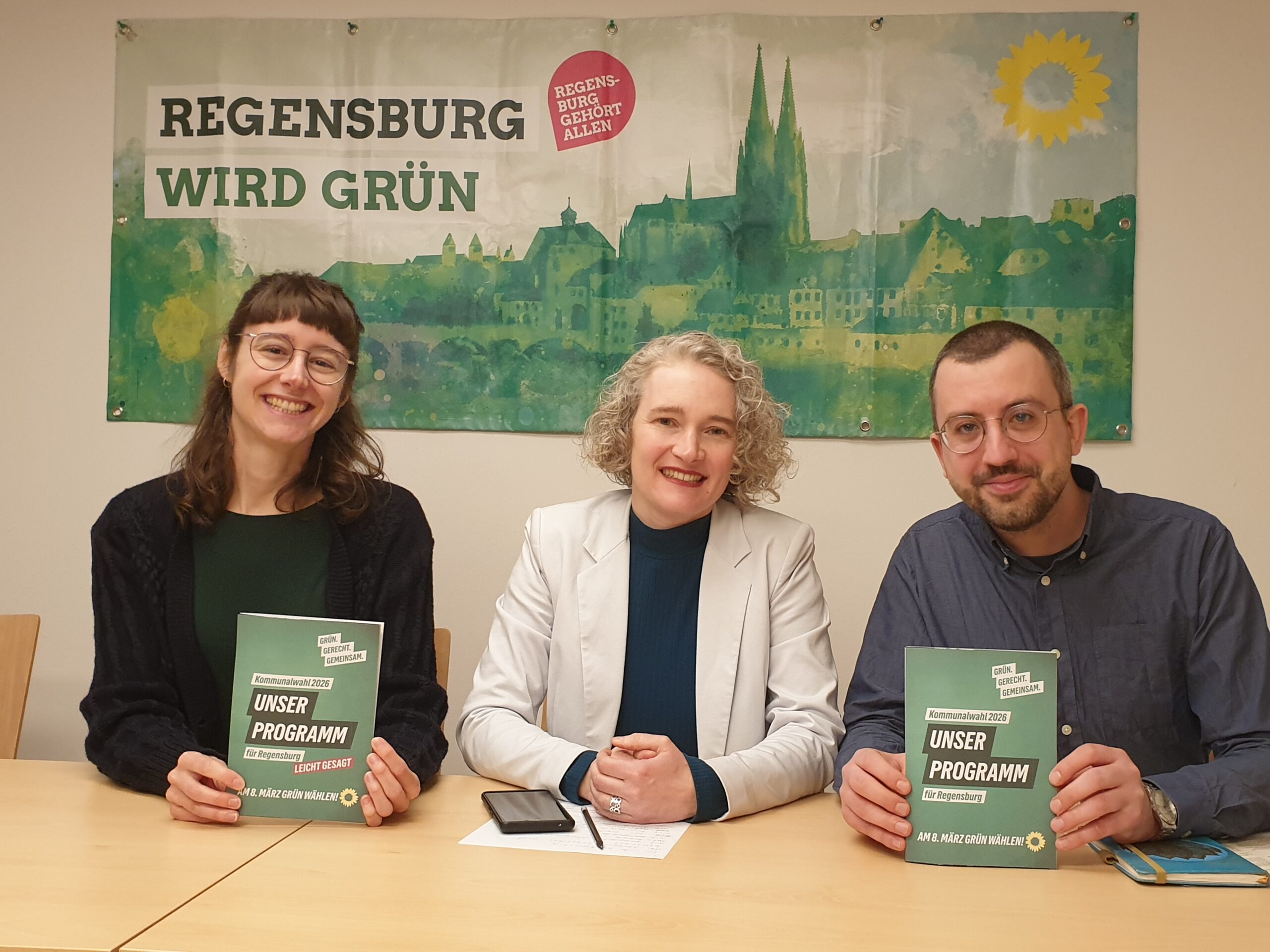 Marie Scholz, OB-Kandidatin Helene Sigloch und Daniel Gaittet stellen das Kommunalwahlprogramm der Regensburger Grünen vor. Sie sitzen nebeneinander vor einem grünen Banner der Regensburger Stadtkulisse und halten die ausgedruckten Cover des Wahlprogramms und des Wahlprogramms in leichterer Sprache in den Händen.