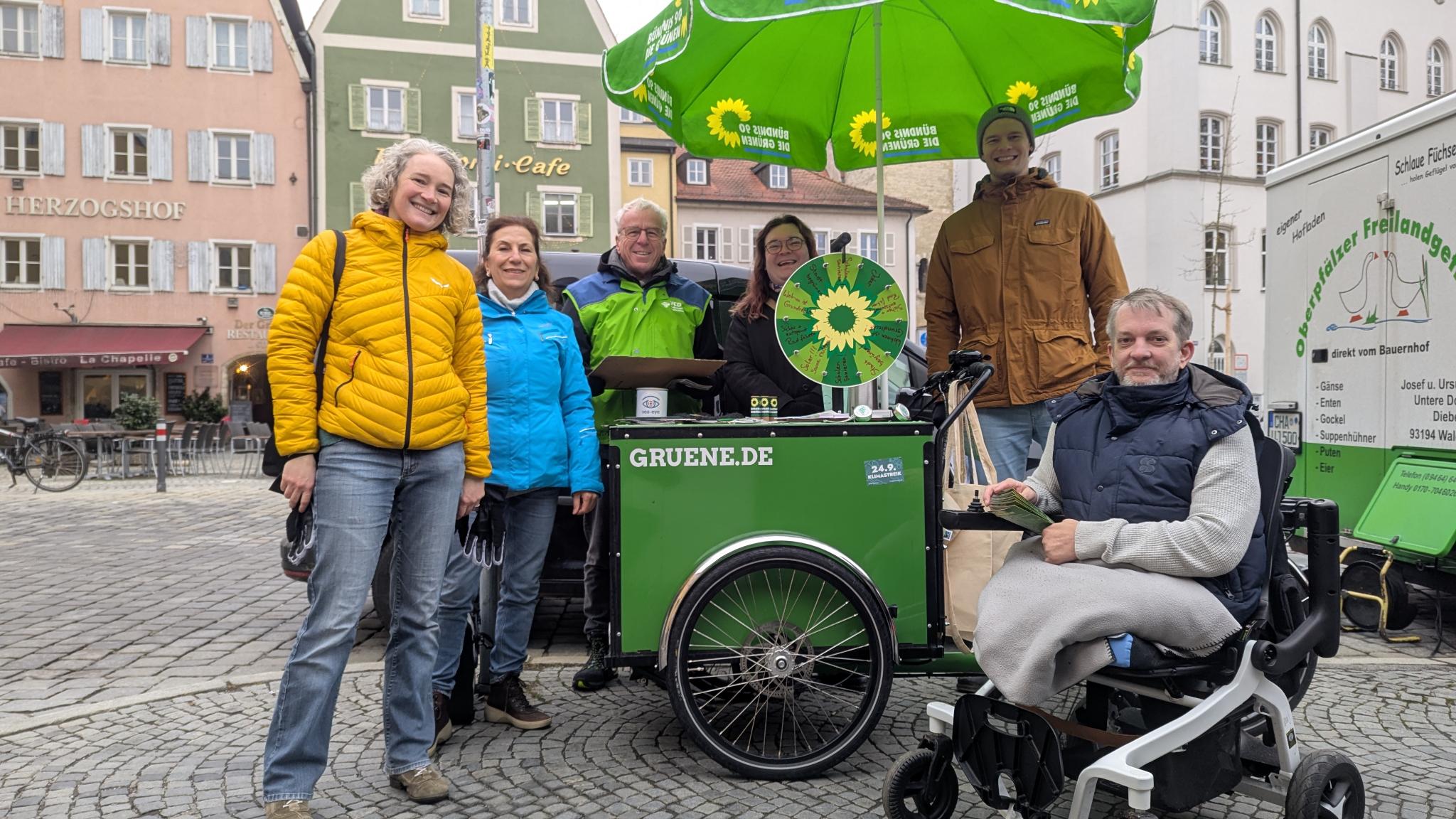 Infostand der Grünen mit grünem Lastenrad, Glücksrad und Sonnenschirm am Kornmarkt. Die Kandidierenden Dr. Helene Sigloch, Monir Shahedi, Wolfgang Bogie, Claire Lindner, Leonhard Stein und Marcel Hylla stehen um diesen und schauen lächelnd in die Kamera.